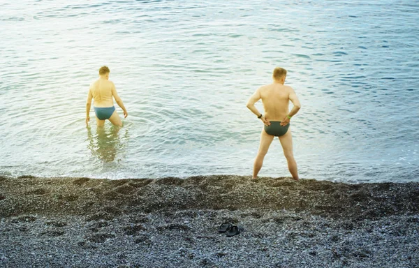 Two adult men stand on a pebble beach and prepare to swim in the cold sea at dawn, back view, sunlight