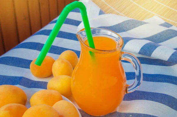 Summer drink-apricot juice in a glass jug with straw and fruit on a striped napkin on a Sunny summer day — Stock Photo, Image