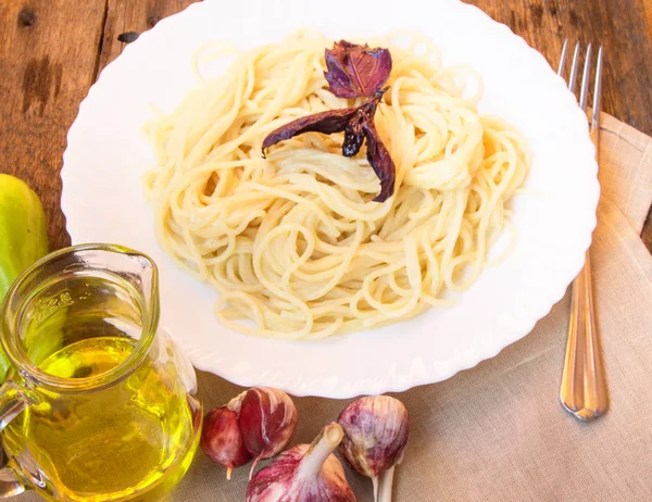 Паста-спагетти в белой тарелке с базиликом на старом деревянном столе с овощами и оливковым маслом в стеклянном кувшине — стоковое фото