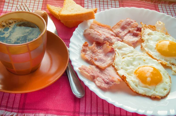 Finom reggeli - egy csésze kávé, egy tányér sült tojás, szalonna és pirítós, mellett az evőeszközök piros kockás szalvéta — Stock Fotó