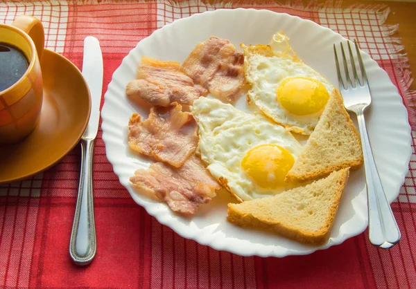 Finom reggeli - egy csésze kávé, egy tányér sült tojás, szalonna és pirítós, mellett az evőeszközök piros kockás szalvéta — Stock Fotó