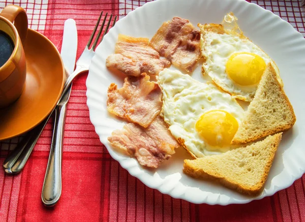 Вкусный завтрак - чашка кофе, тарелка жареных яиц, бекон и тост, рядом со столовыми приборами на красной клетчатой салфетке — стоковое фото