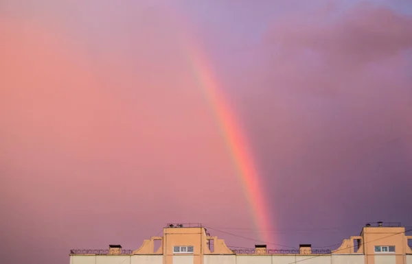 Arco iris sobre el techo de una casa de ciudad de varios pisos en el cielo del atardecer rosado de la noche después de la lluvia, verano fantástico hermoso paisaje mágico — Foto de Stock
