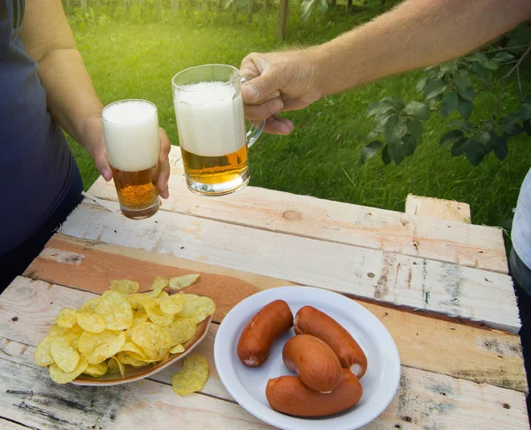 Hands of elderly men and women holding glasses of beer, chips and sausages lie on light wooden boards, people relax on weekends, October festival