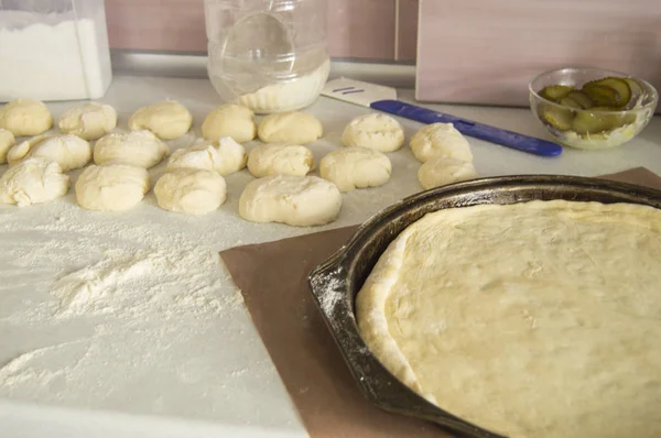 Verse rauwe pizzadeeg op een ronde metalen bakplaat, basis en ingrediënten op een witte keuken tafel bestrooide met meel — Stockfoto