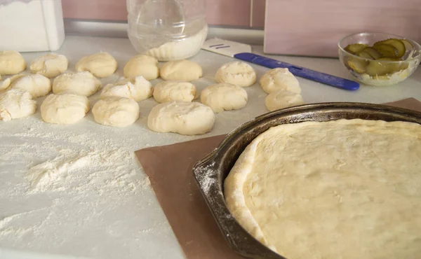 Свежее сырое тесто для пиццы на круглой металлической выпечке, основание и ингредиенты на белом кухонном столе, посыпанном мукой — стоковое фото
