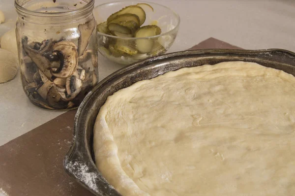 Masa fresca de pizza cruda en una bandeja redonda para hornear de metal, base e ingredientes en una mesa de cocina blanca espolvoreada con harina —  Fotos de Stock