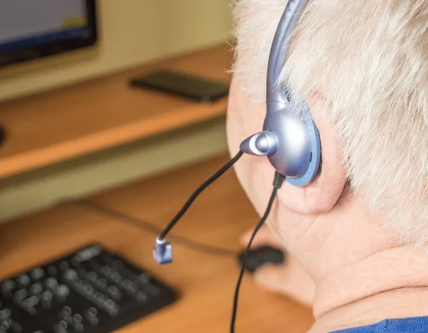 A gray-haired fat old man with a headset looks at the monitor screen, remote work, computer technology for pensioners, adaptation of disabled people to modern life.