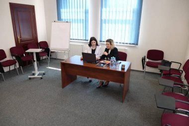 İki kadın konferans odasında dizüstü bilgisayarla çalışıyor, belgeleri inceliyor ve ofis toplantısı sırasında proje stratejisini tartışıyorlar.