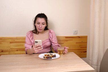 Pembe bluzlu kadın ahşap masada tatlı kek yiyip çay içerken akıllı telefonu kontrol ediyor..