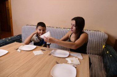 Erkek ve kız kardeş Noel ağacının olduğu bir odaya sofra, tabak, çatal bıçak takımı ve peçeteler kurdular. Ilık ışıklandırma ve yumuşak odaklanma sıcak bir aile ortamı yaratır..