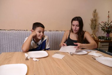 Erkek ve kız kardeş Noel ağacının olduğu bir odaya sofra, tabak, çatal bıçak takımı ve peçeteler kurdular. Ilık ışıklandırma ve yumuşak odaklanma sıcak bir aile ortamı yaratır..