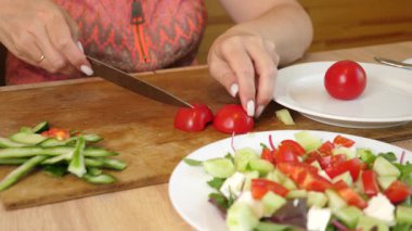 Kadın taze kırmızı domatesleri dilimliyor ve salata tabağına salatalık, yeşillik ve beyaz peynirle ekliyor..