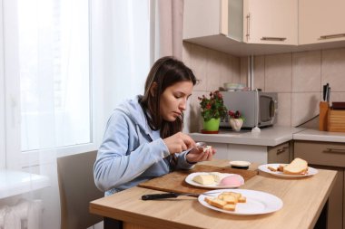 Mutfakta elinde bir tabak sandviçle oturmuş, kaşıkla peynir yiyen kız..
