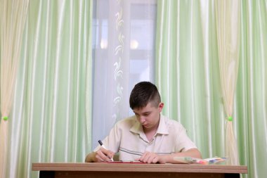 Çocuk masada oturur ve siyah bir kalemle resim çizmeye devam eder..