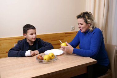 Eli bandajlı çocuk masada otururken mavi kazaklı kadın elinde elma ve bıçakla meyve hazırlıyor..