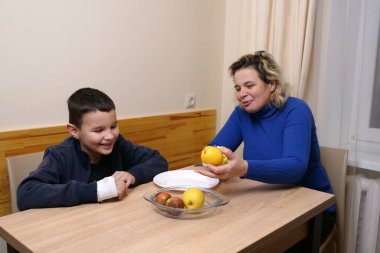 Kadın ona sarı bir elma gösterirken masada gülümseyen çocuk sıcak bir aile ortamı yaratıyor..