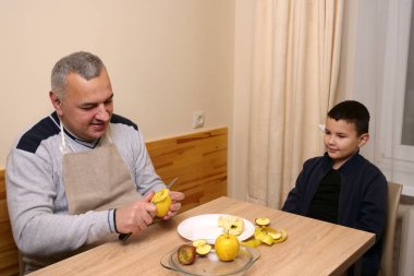 Önlüklü adam bıçaklı elmayı soyarken yanındaki çocuk dikkatle izliyor..