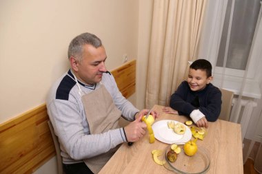 Önlüklü bir adam elmayı bıçakla keserken çocuk gülümseyerek hazırlığı izliyor..