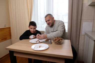Adam temiz ceviz çekirdeğini oğluyla paylaşıyor, çocuk tabağa dikkatlice bakıyor, yakınlarda bir kase ceviz var..