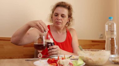 Kırmızı elbiseli kadın mutfak masasında oturuyor, salata yiyor, içecek içiyor, fındık kabuğu soyuyor ve sebze tadıyor..