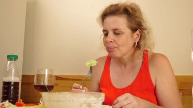 Kırmızı elbiseli kadın mutfak masasında oturuyor, salata yiyor, içecek içiyor, fındık kabuğu soyuyor ve sebze tadıyor..