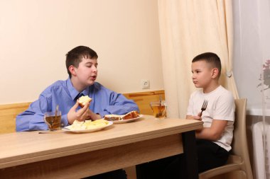 Masada iki çocuk, biri mavi gömlekli, ekmek yiyen, diğeri elinde çatalla dinleyen..