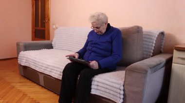 Yaşlı kadın koltukta tablet kullanıyor. Cihazla çalışırken konsantre oluyor..