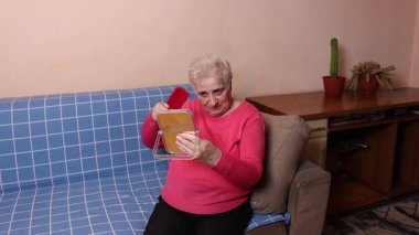 Kırmızı kazaklı yaşlı bir kadın, elinde ayna, saçlarını kırmızı tarak ile tarıyor ve günlük bakım rutinini gösteriyor..