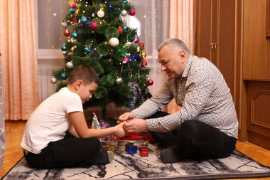 Çocuk ve adam Noel ağacının yanında oturur, küçük hediye süslemelerine bakarlar..