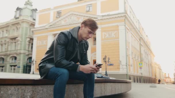 Joven hombre brutal utiliza un teléfono inteligente mientras está sentado en una hermosa ciudad europea . — Vídeo de stock