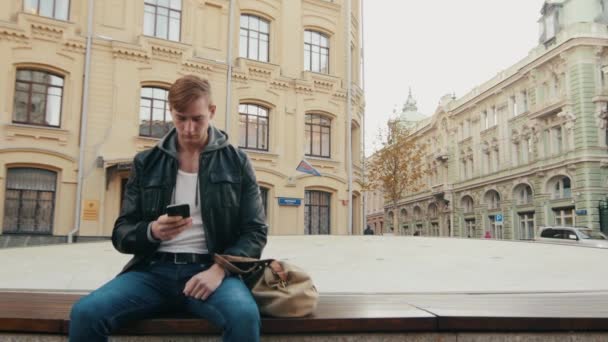 Junger brutaler Mann benutzt ein Smartphone, während er in einer schönen europäischen Stadt sitzt. — Stockvideo