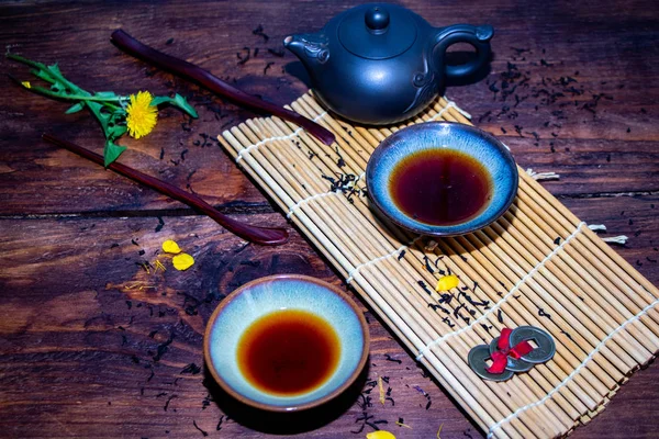 Chinese dishes for the Chinese tea ceremony. Teacups filled with tea, teapot of brown color, wooden objects for tea ceremony. Dry tea is scattered on a brown background.