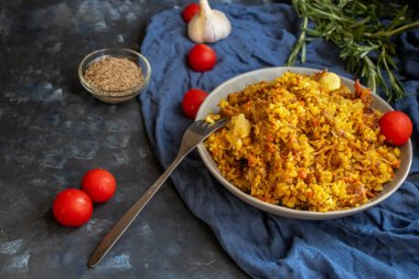 Özbek pilavı yla dolu büyük bir tabak, kiraz domatesin yanında koyu bir bez, elle örülmüş bir standda taze biber. Şeffaf bir tabak, baharat - sıfır ve biber karışımı. Hepsi karanlık bir arka planda.