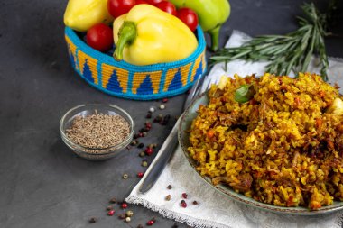 Özbek pilavı yla dolu büyük bir tabak, kiraz domatesin yanında koyu bir bez, elle örülmüş bir standda taze biber. Şeffaf bir tabak, baharat - sıfır ve biber karışımı. Hepsi karanlık bir arka planda.