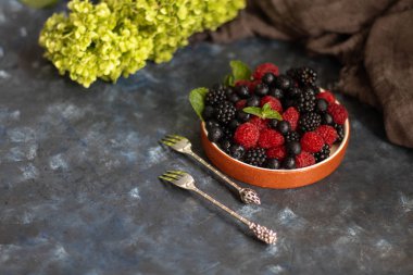 Kahverengi kenarı çilekli bir tabağa koydum. Yaban mersini, ahududu, böğürtlen yan yana, nane yaprakları ile karanlık bir arka plan üzerinde yalan.