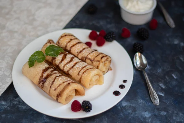 Pancakes with curd filling. Watered with chocolate topping on top, decorated with raspberries and blackberries. In bowls of berries and curd cream.