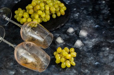 Beyaz üzümler siyah bir tabakta uzanır. Yakınlarda gül şarabıyla dolu iki bardak vardır. Dekorasyon için buz küpleri. Hepsi karanlık bir arka planda.