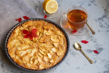 Yuvarlak şekilli elmalı turta, vantilatör katkılı elma dilimleri ile süslenmiş, bir fincan sıcak çay, sonbahar yaprakları, hepsi açık renkli, mermer gri arka plan. 