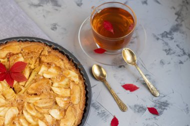 Yuvarlak şekilli elmalı turta, vantilatör katkılı elma dilimleri ile süslenmiş, bir fincan sıcak çay, sonbahar yaprakları, hepsi açık renkli, mermer gri arka plan. 