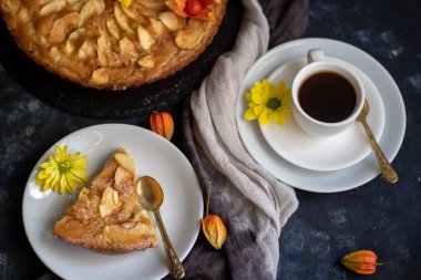 Yuvarlak şekilli Charlotte elmalı turtası, vantilatör yüklü elma dilimleriyle süslenmiş, yanında bir parça kek, bir fincan kahve, sonbahar yaprakları, hepsi koyu bir arka planda.. 