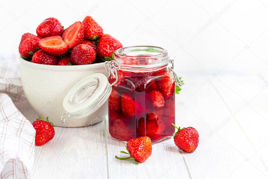 Strawberry jam in a glass jar next to fresh strawberries. On a white wooden background. Homemade winter fruit blanks. Selective focus. Copy space