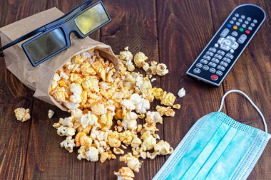 Ahşap bir yüzeyde şeffaf bir kase patlamış mısır. Laptop ve kulaklığın yanında. Maske virüslere karşı korur. Boşluğu kopyala.