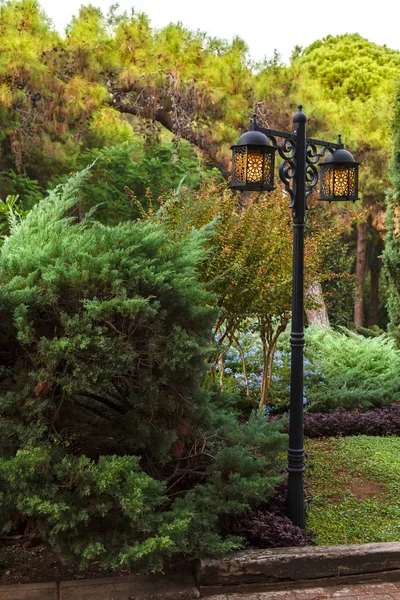 Beautiful metal street lamp with yellow lamps on a background of green trees and bushes on a sunny day