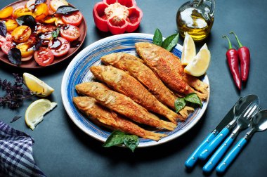 Kızarmış deniz ürünleri ('red mullet', 'barbule') üzerinde mavi bir plaka, koyu arka plan üzerinde. Sonraki plaka sebze salatası ve kırmızı biber ile