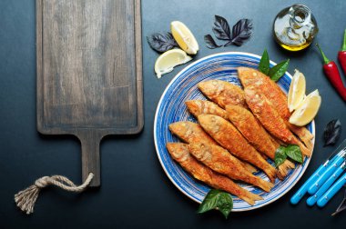 Kızarmış deniz ürünleri ('red mullet', 'barbule') üzerinde mavi bir plaka, koyu arka plan üzerinde. Sonraki kesme tahtası