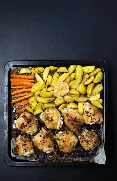 Ready meal. Chicken thighs, potatoes, carrots and onions baked on a baking sheet. Dark background. Place for text