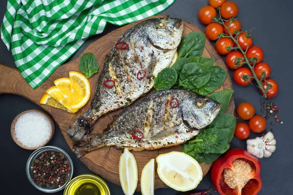 Dorado pescado al horno en especias sobre una tabla de madera marrón. Cerca de espinacas, limón, aceite de oliva, tomates cherry, pimienta — Foto de Stock