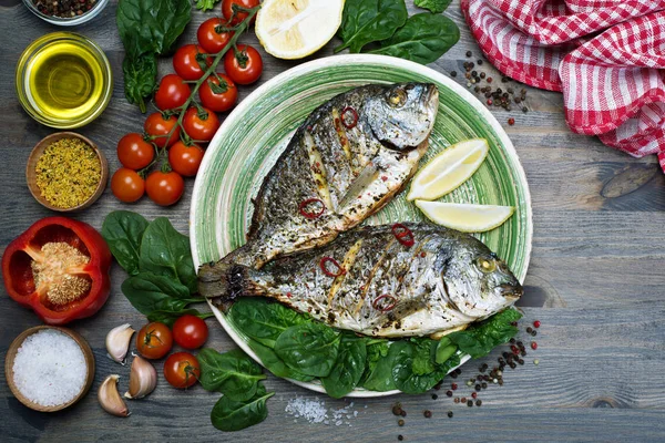 Рыба Дорадо запекается в духовке в специях на зеленой тарелке на сером деревянном фоне. Рядом шпинат, лимон, оливковое масло, помидоры черри, перец — стоковое фото