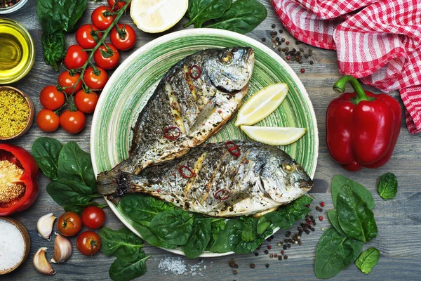 Рыба Дорадо запекается в духовке в специях на зеленой тарелке на сером деревянном фоне. Рядом шпинат, лимон, оливковое масло, помидоры черри, перец — стоковое фото